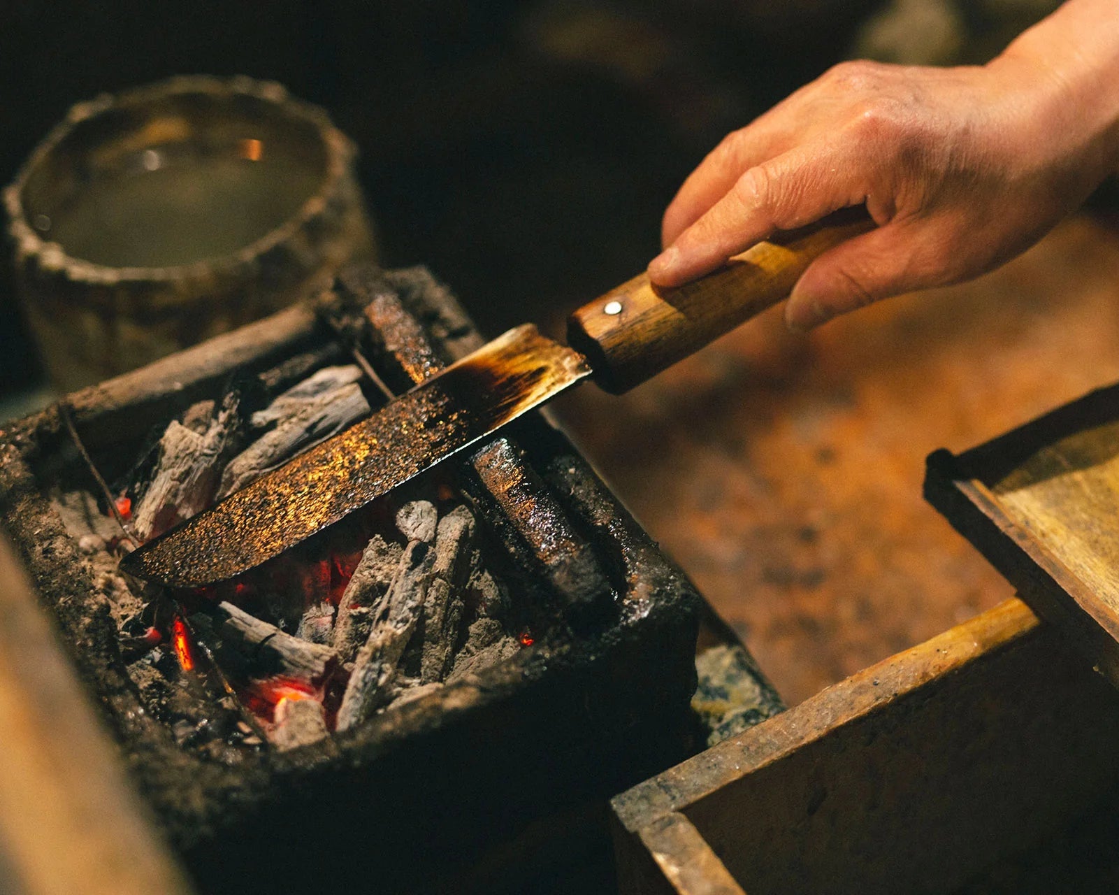 Hand holding a metal spatula over a fire pit with glowing embers for Japanese candle making.
