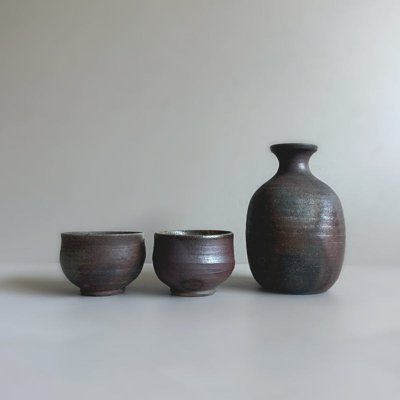 a handcrafted Tsugaru Kanayama pottery sake set made in Japan, featuring a dark earthy brown finish. The set includes two sake cups and a traditional sake bottle.