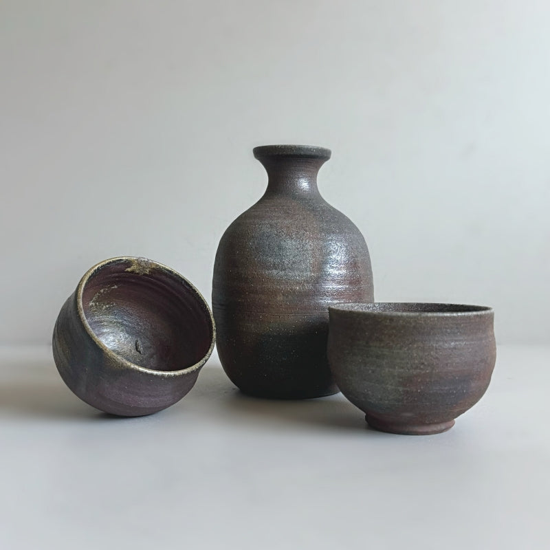 a handcrafted Tsugaru Kanayama pottery sake set made in Japan, featuring a dark earthy brown finish. The set includes two sake cups and a traditional sake bottle.